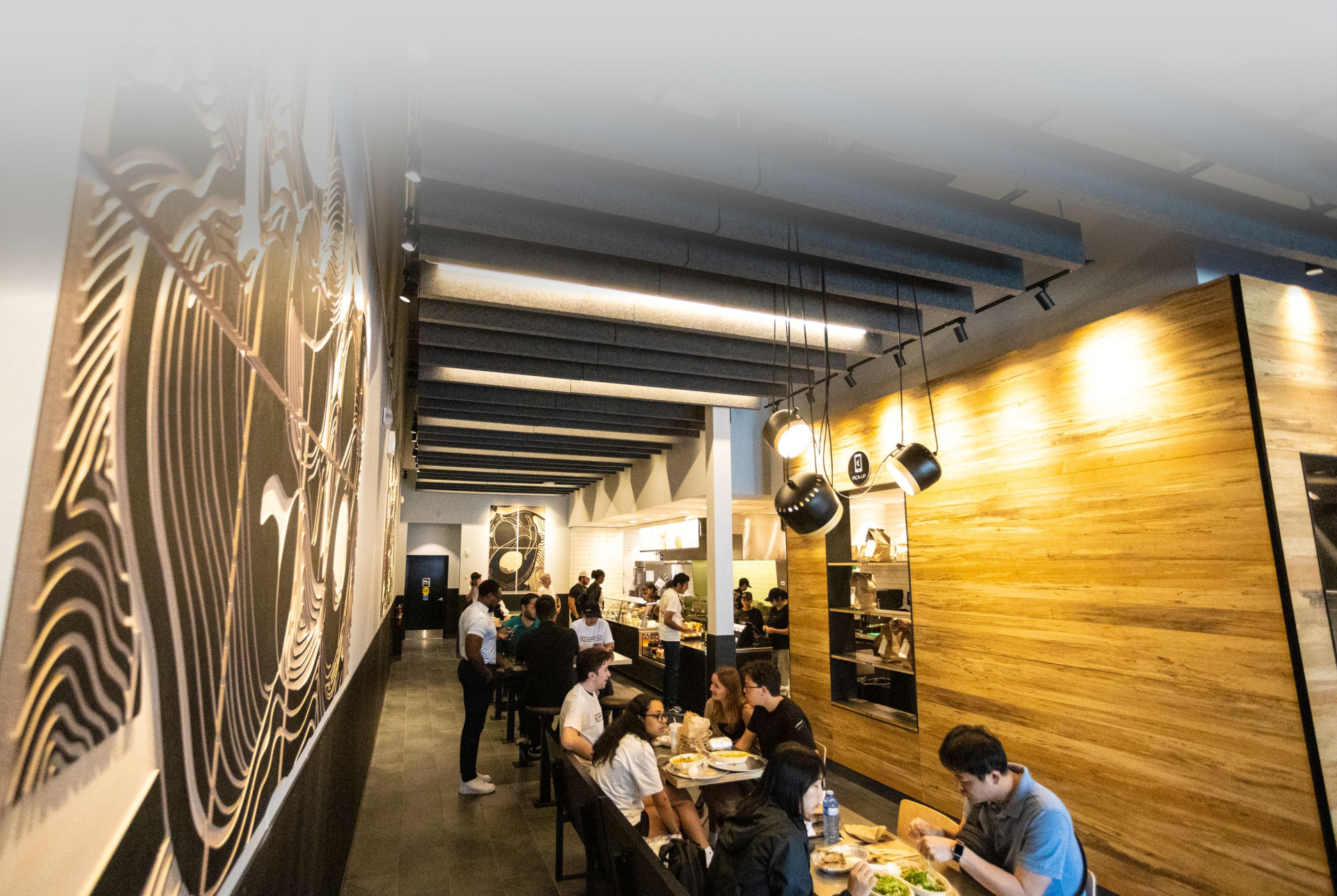 A busy restaurant interior with long tables and bench seating, where patrons are dining along a narrow aisle. Wood-paneled walls, exposed ceiling beams, and hanging lights create a warm, modern atmosphere.